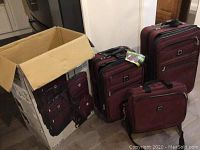 Photo showing the three pieces of burgundy luggage standing next to the original packaging box depicting the images of the luggage set.