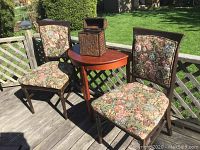 Two floral brocade upholstered wood-framed dining chairs, reddish-brown wooden demilune table, and rectangular woven wicker box on a wooden deck outdoors under sunlight.