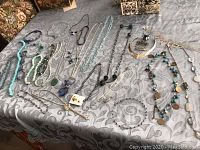 Wide view of approximately 25 costume jewellery necklaces laid out on tablecloth, showing variety in style, beads, and metal chains.