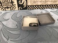 Ring in gray box with soft white padding inside, shown from angle highlighting the band surface and edge pattern.