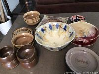 Full view of the 16 assorted pottery pieces on a tabletop, including bowls, plates, and jars. Shows variations in size and glaze, brown and blue accents visible.