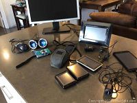 Wide view of all electronic items on table showing HP tablet, Vivitar camera in case, ViewSonic digital frame, Dell monitor, computer mouse, GPS units, and blue Bud Light mini speakers.