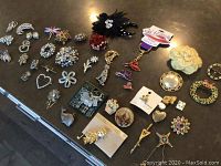 Top view of approximately 35 assorted brooches and pins laid out on table showing variety of designs, colors, and styles including floral, hearts, stars, and rhinestone accented pieces.