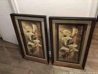 Two framed calla lily flower prints side by side on floor against a white wall, showing front view of prints in brown frames.