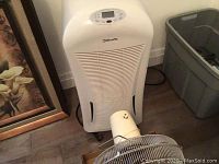 Photo of white Danby Silhouette dehumidifier with front vents and top control panel, positioned on floor next to a grey bin and framed picture.