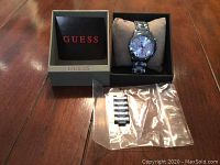 Guess men's silver-tone watch on a beige cushion inside the black Guess watch box. Extra metal links in a sealed plastic bag placed next to the box. Outer grey sleeve with Guess branding visible.