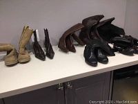 Seven pairs of various boots displayed on a counter; styles include suede winter boots, leather ankle and mid-calf boots in brown and black colors.