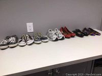 Six pairs of shoes lined up next to each other on a white surface against a gray wall. Includes Converse, Adidas, Tod's, and Reef shoes in various colors and styles.