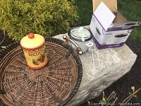 Overall lot contents showing wicker tray holding biscotti jar, rice cooker and box on stone