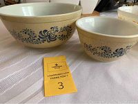 Two vintage Pyrex Homestead mixing bowls on a white tablecloth with blue floral design on cream speckled background.