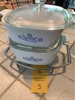 Photo showing two Corning Ware Cornflower Blue casseroles stacked on metal trivet with glass covers, highlighting pattern and condition
