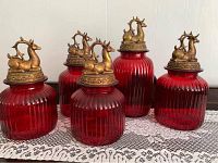 Set of 5 red ribbed glass canisters each topped with brass lids decorated with brass reindeer figures. Displayed on a lace cloth.