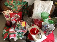 Overview of Christmas-themed holiday table linens, pot holders, mugs, glassware, candles, and pewter salt and pepper shakers arranged on chairs and table.