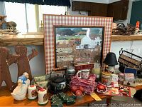Wide view of various country cottage-themed decorative and gift items arranged on a counter, including the framed print, wooden gingerbread men, ceramic duck figurine, kettle, kitchen jars, and fabric items.