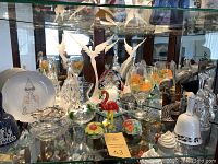 Wide shot of the curio cabinet shelf showing the Lavaggi Soaring Angel figurine, glass bells, technical glasses, flamingo figurine, and various small glass items.
