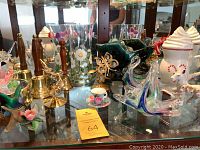 Photo showing a display of glass figurines including birds, porcelain cup, green glass bowl, white ceramic container with candy cane decoration, brass bells with wooden handles