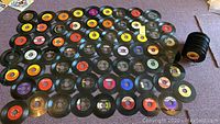 Image showing over 60 vinyl records spread out flat on a carpeted surface, most appear 7 inch, no covers.