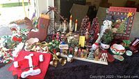 Wide view of the Christmas and winter decor items arranged on table, includes Santa clothing, gold bells, assorted figurines, mugs, and glassware