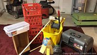 Wide view of mop bucket, crates, brooms, and container on red carpet.