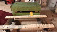 Green wooden box platform utility cart with two wheels and visible wear marks, photographed with wooden slat platform cart beside it and two white painted wooden planks in front for size reference.