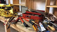 Vintage red metal tool box and various hand tools including cutters, saw, wrenches, screwdrivers, and yellow extension cord displayed on wooden shelves.