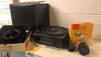 Kodak 750H Carousel projector with accessories including lens, cardboard box for slide loader, and carrying case.