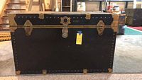 Front view of vintage trunk showing black body, brass corner protectors and central lock.