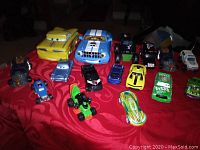 Photo showing 16 toy cars arranged wide on red fabric surface, including various types and colors such as cartoon-style cars, race cars with numbers, and open-wheel green race car.