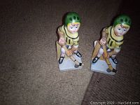 Two ceramic hockey player figurines with green helmets and yellow jerseys number 11, each holding a hockey stick, placed on carpet and photographed from above.