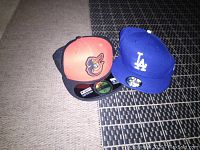 Two New Era baseball caps on a rug; one blue Los Angeles Dodgers cap and one black/orange Baltimore Orioles cap.
