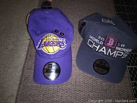 Two caps side by side on a brown surface. Left cap purple with Los Angeles Lakers logo. Right cap dark gray with 2018 World Series Champs Boston Red Sox logo.