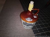 Photo showing ceramic Evan Williams whiskey jug with brown top and cream body, standing on a carpet with a visible cork sealed with yellow wax at the neck.