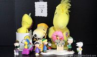 Wide shot showing all 12 Peanuts collectibles arranged in front of a black backdrop. Includes plush toys, resin figurines, mugs, and wind-up toys.