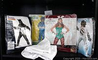 Photo showing all five costumes together on a shelf: two werewolf costume packages, one Cookie Monster costume package, one Playboy Bunny costume package, and one folded white lab coat with tags in front.