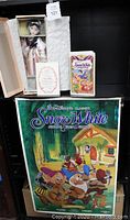 Photo showing the porcelain Snow White doll in original box along with certificate of authenticity and 1994 VHS tape, and rolled poster underneath.