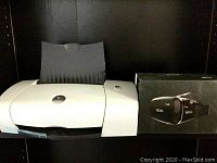 Photo showing white and gray Dell printer with paper tray and VR glasses in box beside it on a shelf.