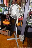 Full view of three electric fans. One is a height adjustable pedestal fan with white plastic blades and protective grill. Two smaller fans are visible on the floor behind the pedestal fan.