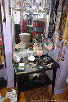 View of the wood table with various makeup mirrors, jewelry including earrings, a small decorative vase, and makeup sponges sitting on the table. The large wood framed mirror hangs above the table reflecting surrounding items.
