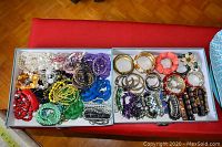 Two trays side by side displaying various plastic bead and metal cuff style bracelets arranged by color and style.