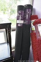Four rolled black area rugs standing vertically near window, two are wrapped with Life At Home label.