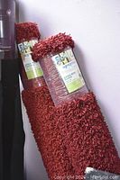 Two rolled red shag style area rugs standing against a wall, showing thickness and texture.