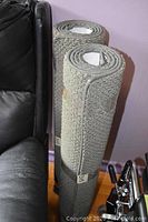 Two rolled gray rugs with tight loopy pile leaning against a black leather sofa.