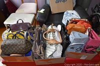 Overall view of at least 15 handbags and purses arranged in boxes, showing variety of colors and styles.