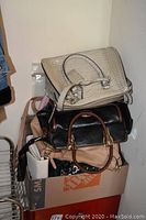 Stack of assorted purses including a large light beige snakeskin embossed purse on top, black purse below, and beige purses underneath in cardboard box.