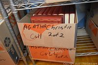 Two cardboard boxes labeled as Agatha Christie collection on metal shelving, showing red hardcover books inside.