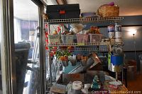 Photo showing wire shelving with various baskets containing glue, scissors, crayons, enamel paint cans, tapes, and craft tools arranged on shelves in a room with window light.