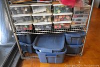 Overview of metal shelving unit with nine plastic containers of craft paints on top shelf and three large blue plastic totes underneath.