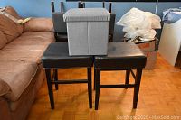 Two black leather padded stools with wooden legs, stacked with a gray fabric storage box on top, against a brown couch and blue wall.