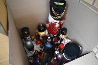 Box full of multiple nutcracker figurines, viewed from above showing variety of sizes and designs, with cloth tote containing ribbons and wrapping paper beside them