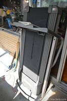 Full folded treadmill, black running belt, gray frame, shown outside leaning against a glass door